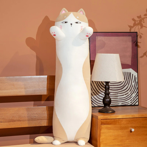 På en säng med ett nattduksbord bredvid, med en vit sänglampa och en ram bakom, ligger en vit och brun kattkudde som står på sina små bakben